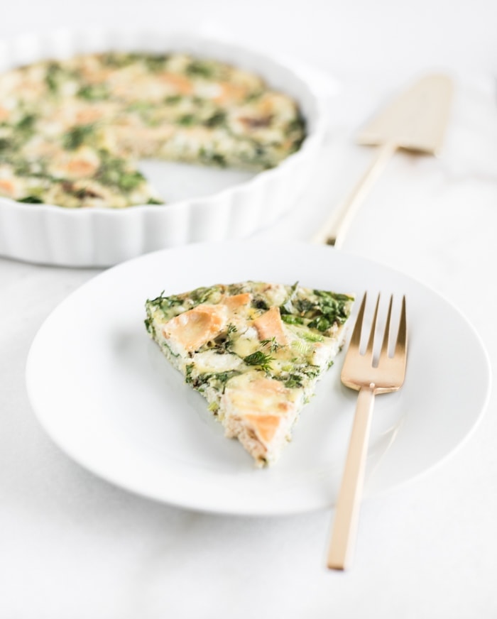 closeup view of a slice of smoked salmon and kale frittata on a white plate with a gold fork.