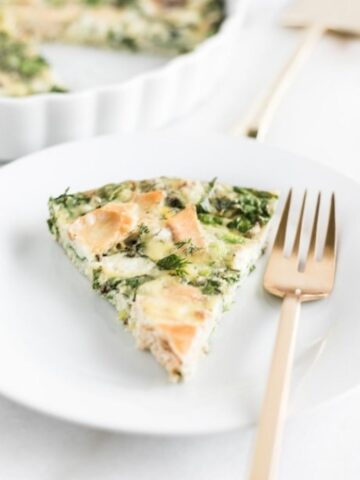 slice of smoked salmon kale frittata in on a white plate with a gold fork next to it.