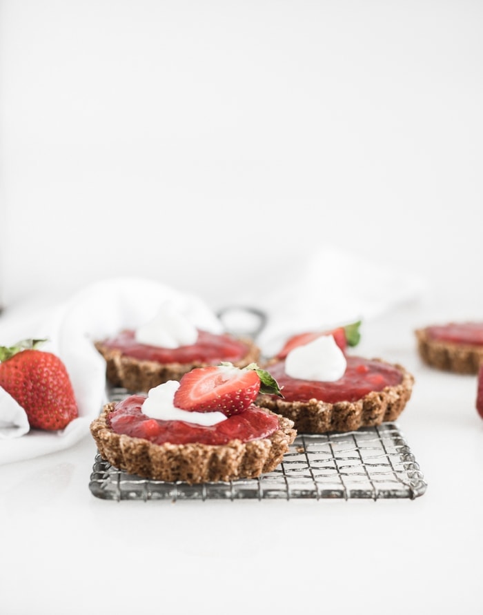 mini strawberry rhubarb tarts with whipped cream and half a strawberry on top on a cooling rack.