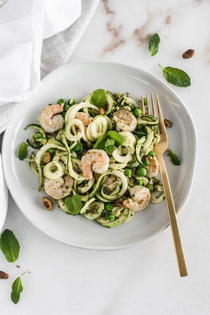 overhead view of zucchini noddles with shrimp and mint pesto on a white plate with a gold fork.