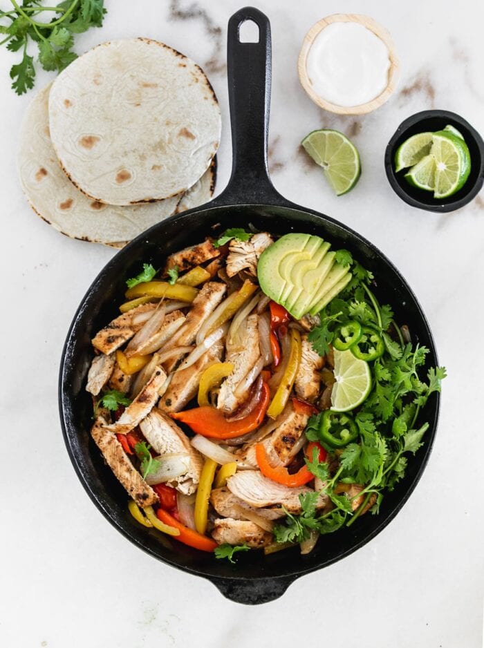 overhead view of easy grilled chicken fajitas on a skillet with bell peppers, cilantro, lime, and avocado.