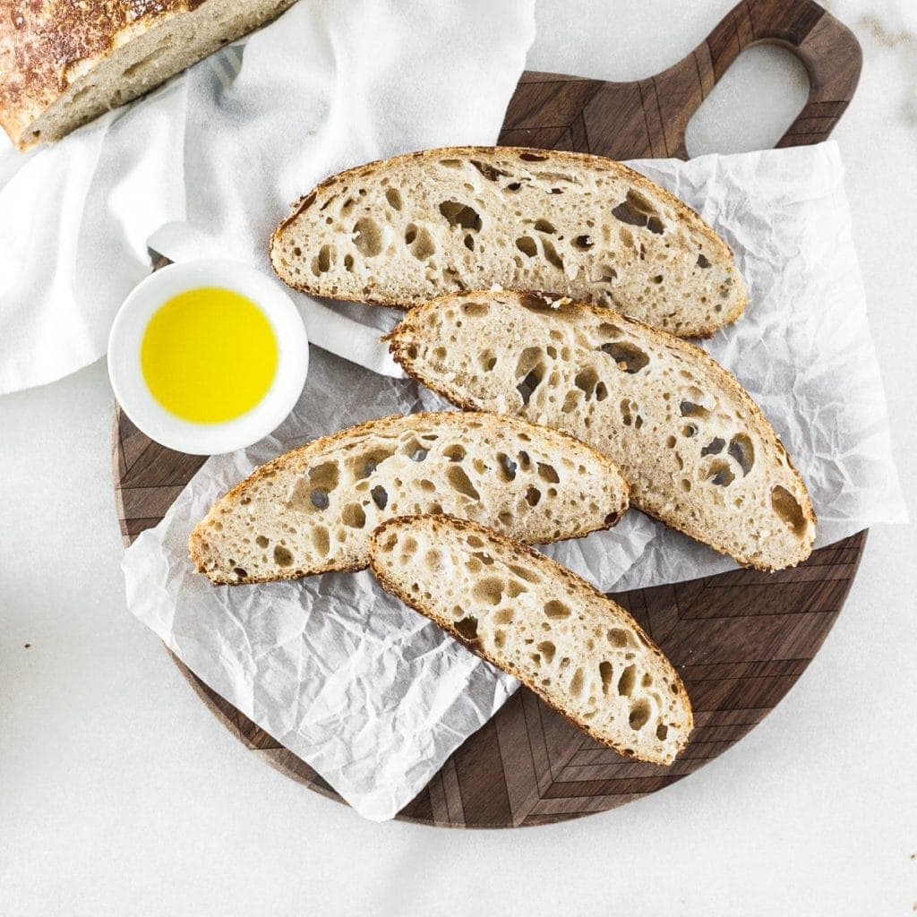 slices of homemade sourdough bread on a round wooden cutting board with a bowl of olive oil. 