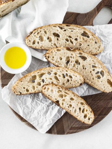 slices of homemade sourdough bread on a round wooden cutting board with a bowl of olive oil.