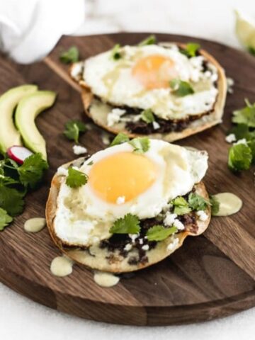 two healthy huevos rancheros tostadas on a wooden serving tray with avocado slices, cilantro and lime wedges.