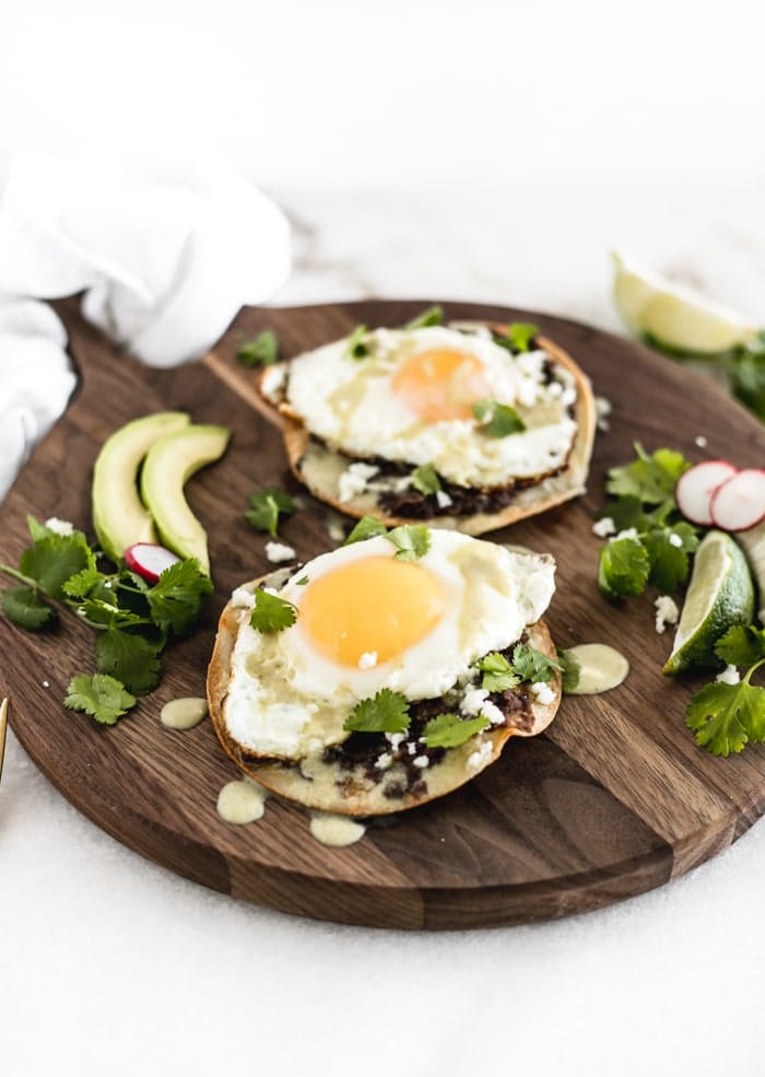healthy huevos rancheros tostadas on a wooden serving tray with extra ingredients around it.