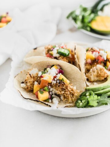 crunchy coconut fish tacos topped with a fresh mango salsa on a white plate.