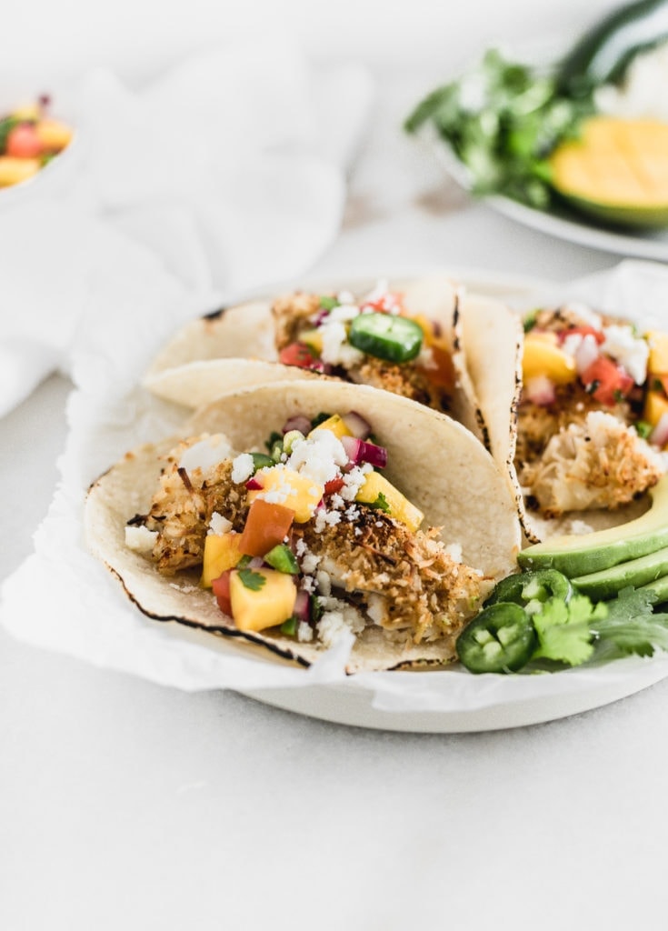 Crunchy coconut fish tacos topped with a fresh mango salsa on a white plate.