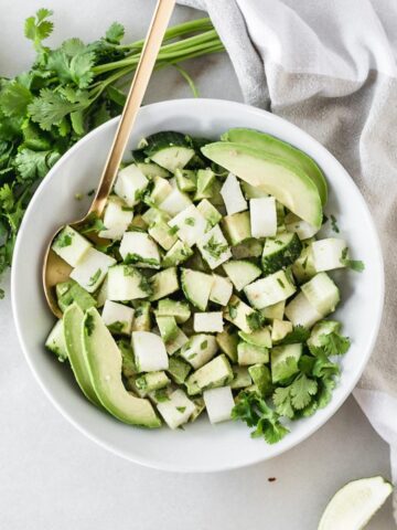 overhead view of Avocado, Jicama and Cucumber Salad in a white bowl with a gold spoon in it surrounded by cilantro. lime wedges and a grey and white linen.