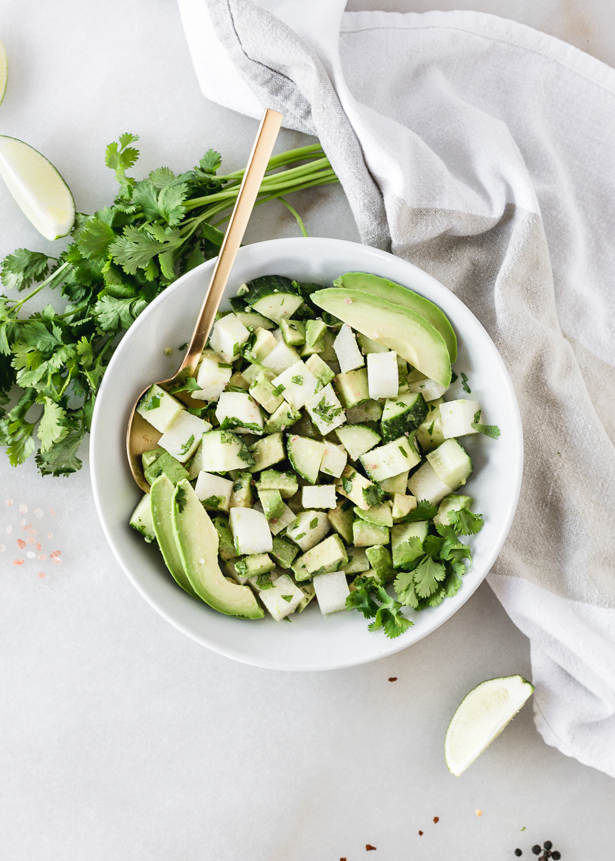 overhead view of avocado jicama cucumber salad in a white bowl with a gold spoon in it surrounded by cilantro, lime wedges and a white napkin.