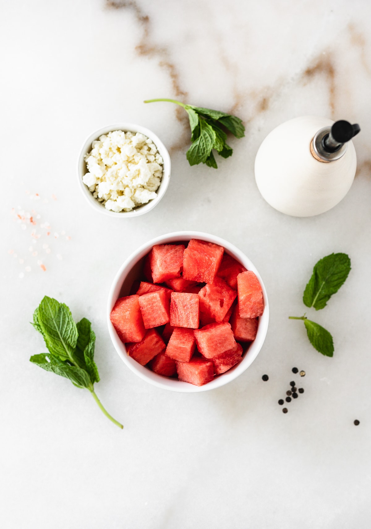 overhead view of a bowl of watermelon. a bowl of feta. mint leaves, and a bottle of olive oil on a white background.