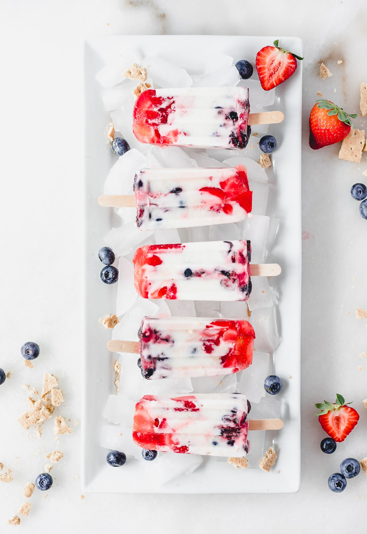 overhead view of red white and blue berry cheesecake popsicles on a platter with ice, berries and crushed graham crackers.