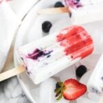 overhead view of red white and blue berry cheesecake popsicles on a platter with ice, berries and crushed graham crackers.