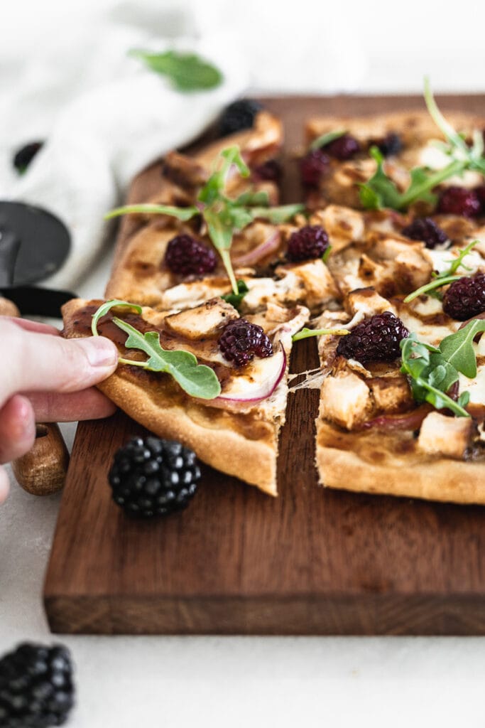 hand taking a corner piece of blackberry chicken flatbread pizza from a cutting board.