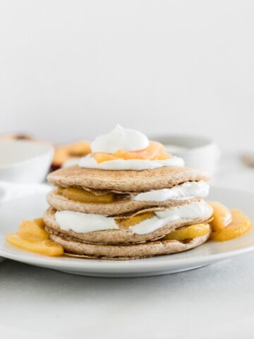 stack of healthy peaches and cream pancakes layered with yogurt and peach compote on a white plate.