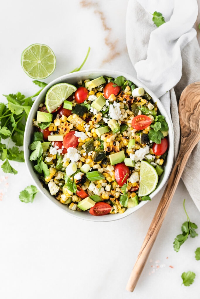 overhead view of grilled corn and poblano salad in a bowl garnished with cilantro and lime wedges with a wooden spoon and white napkin next to it.