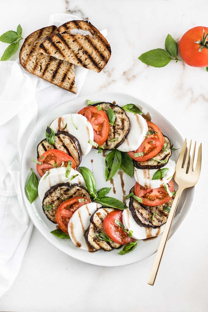 overhead shot of grilled eggplant caprese salad on a plate with a gold fork, surrounded by grilled toast and a tomato.