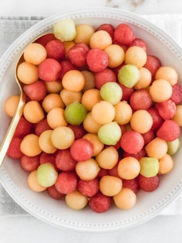 overhead view of melon balls in a white bowl with a gold spoon in it.
