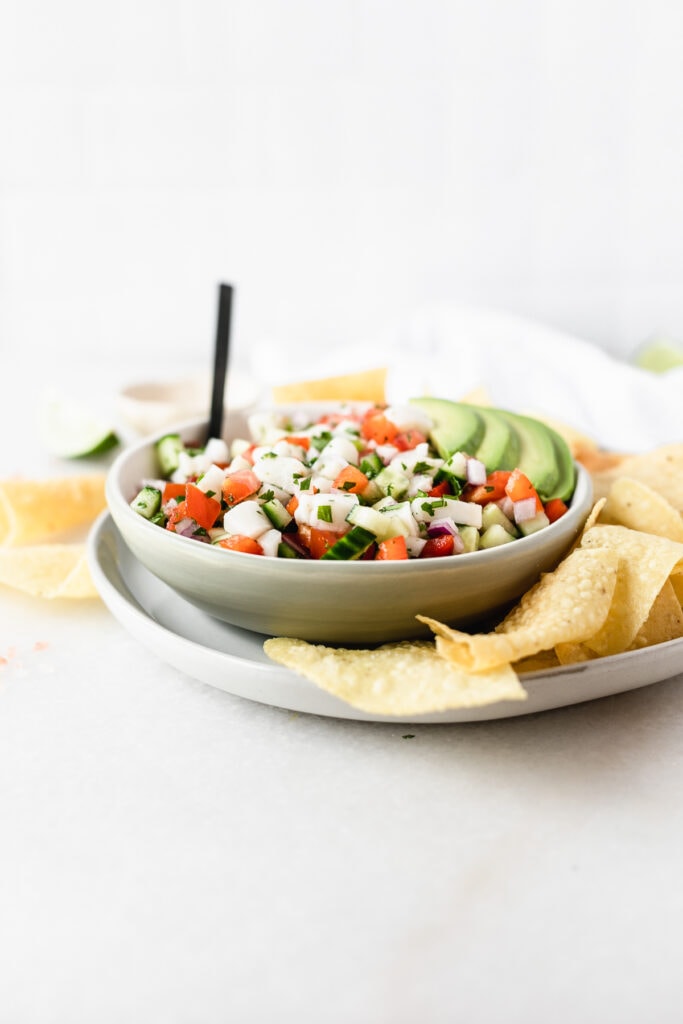 bowl of ceviche with a black spoon in it and sliced avocado on top on a plate with tortilla chips.