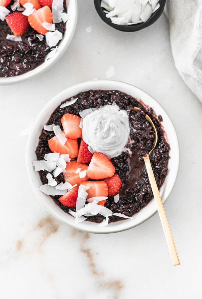 overhead view of black rice breakfast porridge in a bowl with a gold spoon, topped with strawberries, coconut flakes and coconut whipped cream.