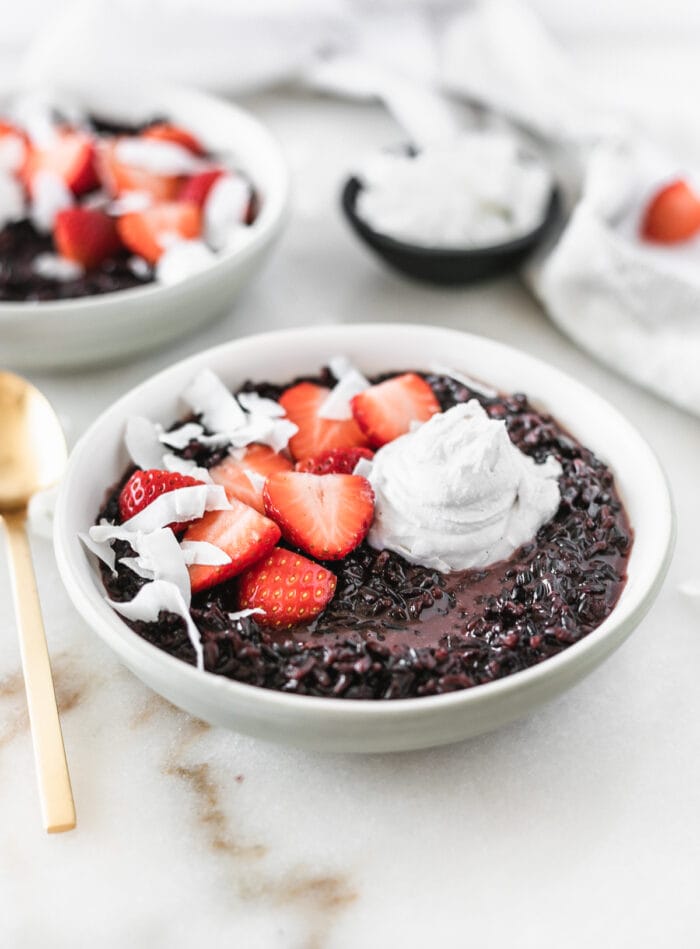 coconut black rice breakfast porridge in a grey bowl topped with strawberries, coconut and coconut whipped cream.