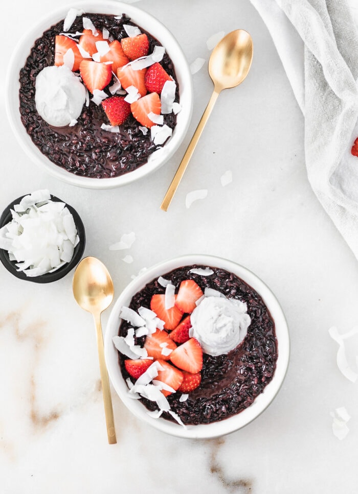 overhead view of two bowls of coconut black rice breakfast porridge topped with strawberries, coconut flakes and coconut whipped cream.