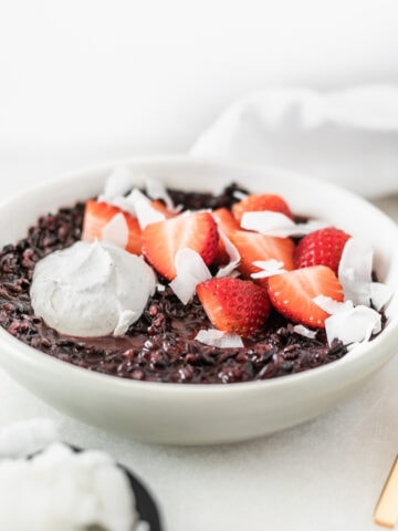 coconut black rice breakfast porridge in a grey bowl topped with strawberries, coconut and coconut whipped cream.