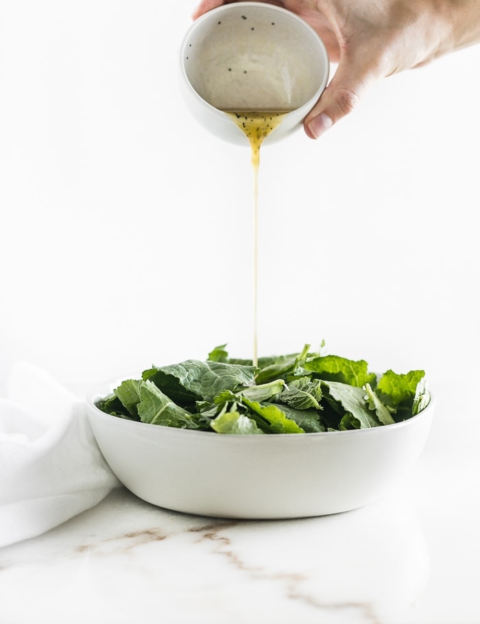 grey bowl with baby kale and a bowl of lemon dressing being poured into it.