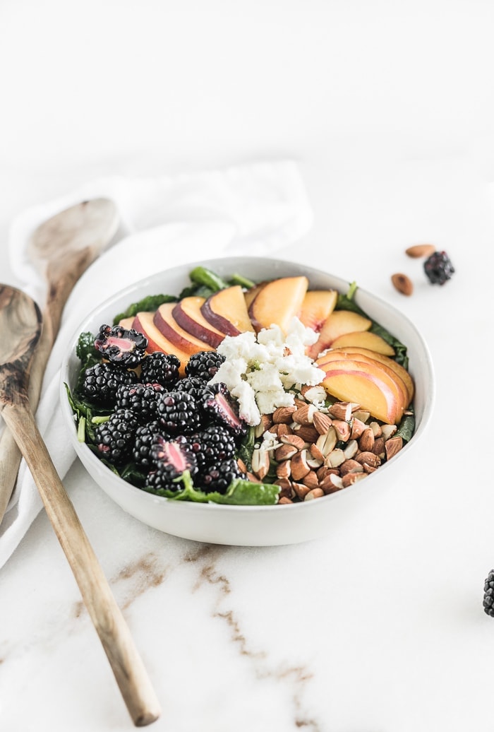 bowl of kale with blackberries, sliced peaches, goat cheese and almonds arranged on top.