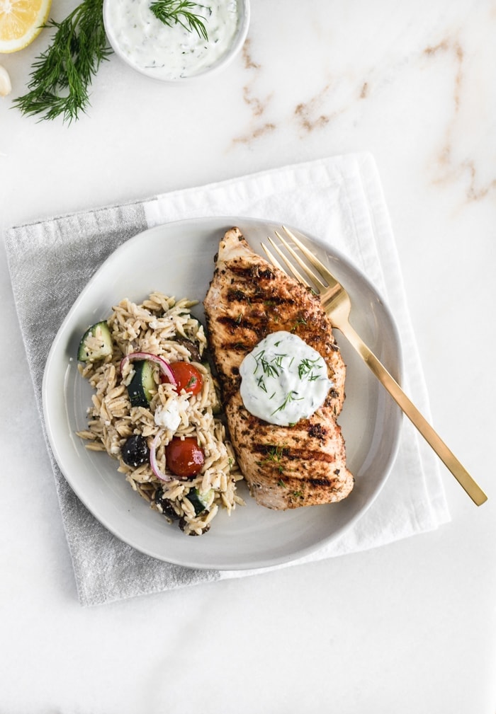 overhead view of healthy Greek chicken with tzatziki sauce on top and pasta salad next to it on a white plate.