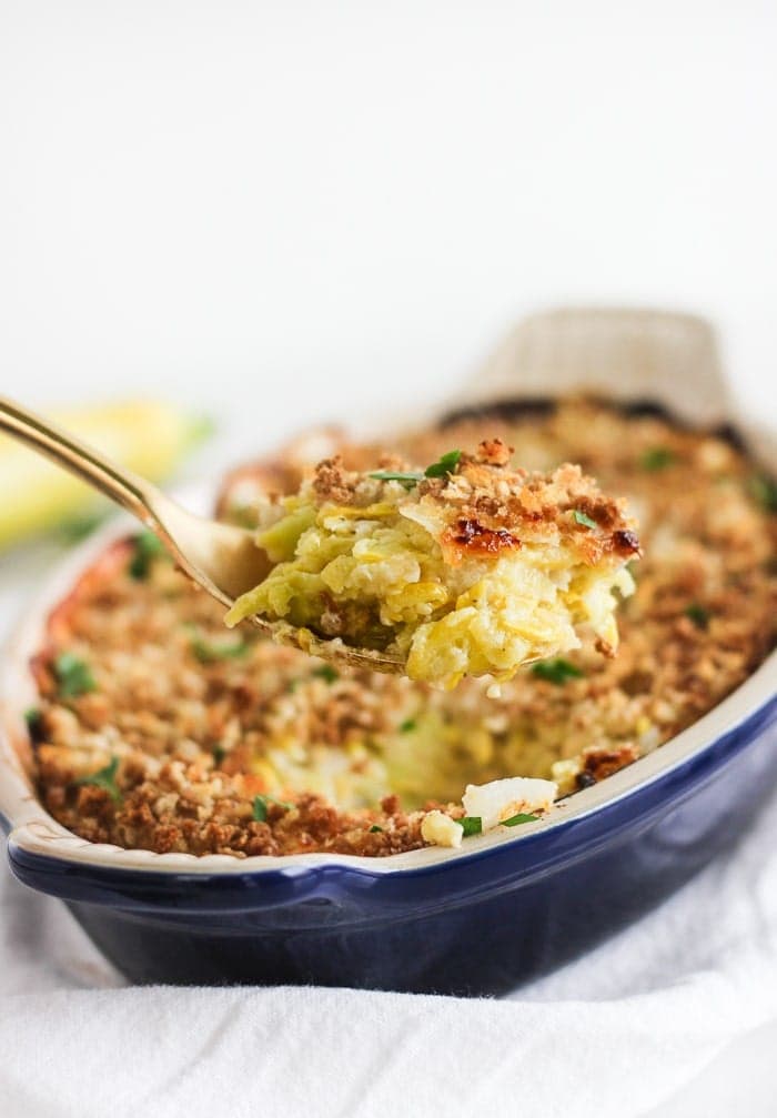 gold spoon lifting cheesy summer squash casserole out of a blue baking dish.