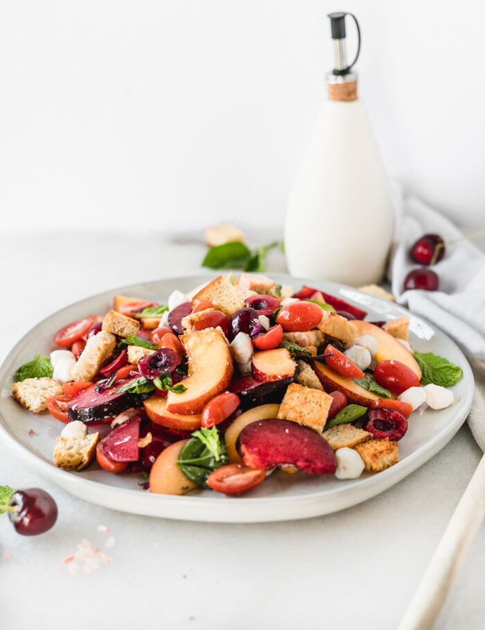 stone fruit panzanella salad on a grey plate with a bottle of olive oil and fruit in the background.