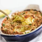 gold spoon lifting cheesy summer squash casserole out of a blue baking dish.
