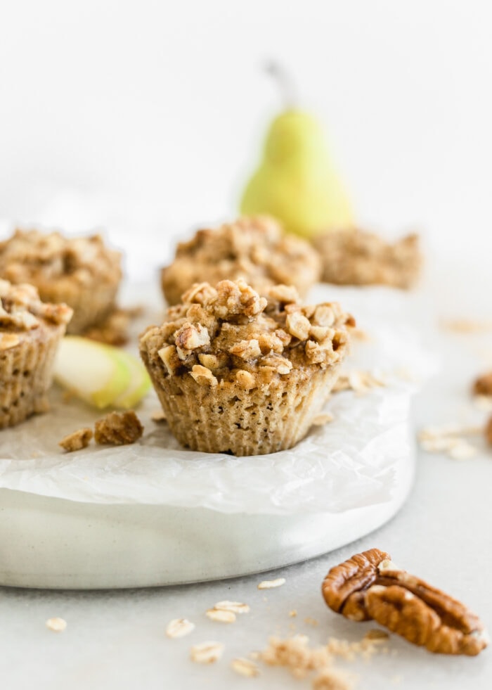closeup of a pecan cardamom pear muffin on a parchment lined plate surrounded by pecans, oats, pears, and more muffins.