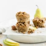 two pear pecan streusel muffins stacked on top of each other on a plate surrounded by streusel topping, pecans, oats and pears.