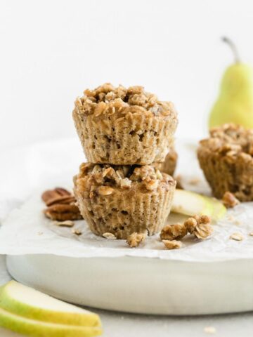 two pear pecan streusel muffins stacked on top of each other on a plate surrounded by streusel topping, pecans, oats and pears.