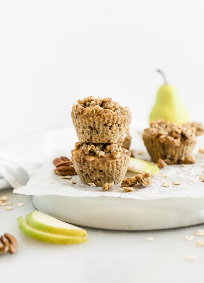 two pecan cardamom pear muffins stacked on top of each other surrounded by pear slices and streusel topping.