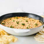 healthier red pepper hot crab dip in a white skillet with chips and crackers around it.