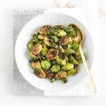 overhead view of sweet chili brussels sprouts in a white plate on top of a gray and white napkin with a gold spoon.