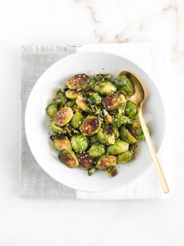 overhead view of sweet chili brussels sprouts in a white plate on top of a gray and white napkin with a gold spoon.