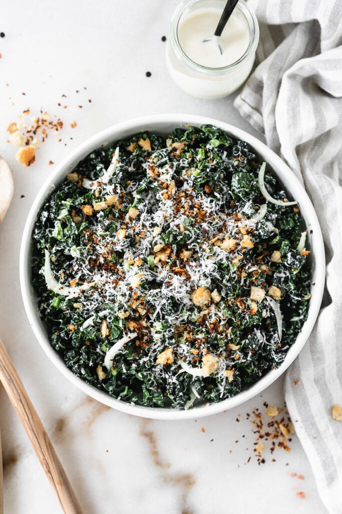 overhead view of kale caesar salad in a white bowl wooden spoons, a striped towel, and a jar of dressing beside it..