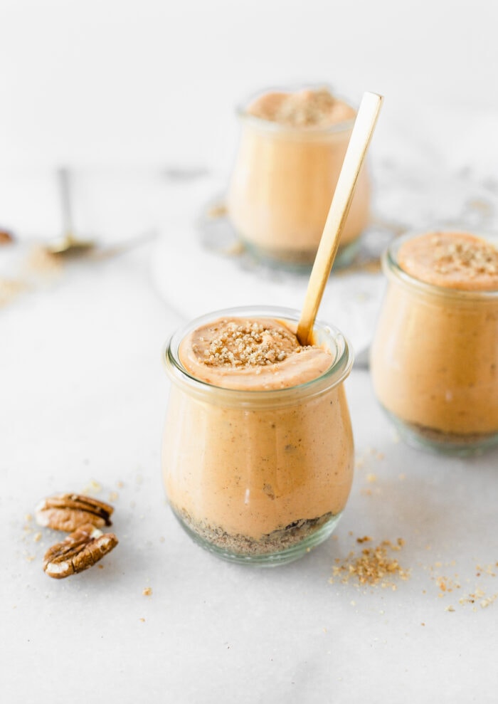 jar of healthier no bake pumpkin cheesecake with a gold spoon in it and two more jars of pumpkin cheesecake in the background.