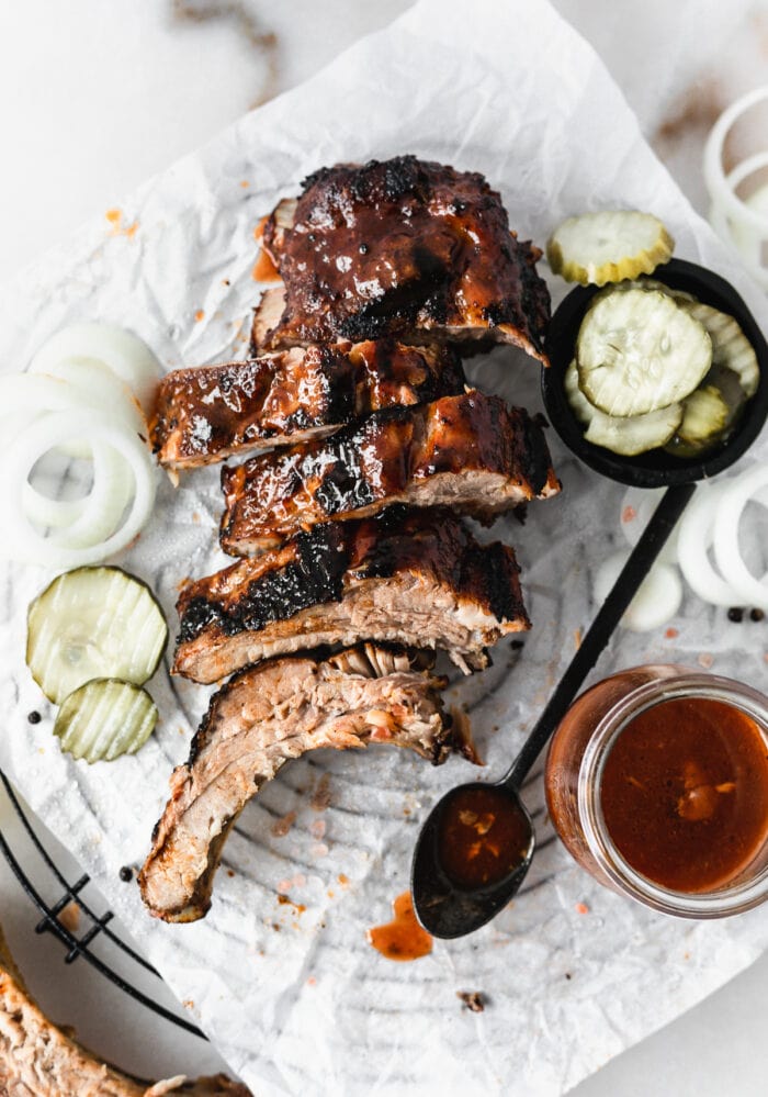 overhead view of sliced bbq ribs on a parchment lined rack with a spoon and jar of bbq sauce, pickles, and sliced onions.