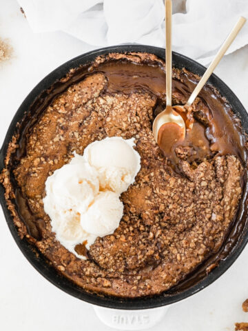 pumpkin lava cake with walnut topping and ice cream on top in a white cast iron skillet with two gold spoons in it.