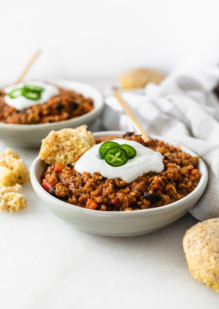 vegan pumpkin lentil chili in a small grey bowl with sour cream, jalapeño slices and cornbread on top.