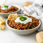 vegan pumpkin lentil chili in a small bowl topped with sour cream, jalapeno slices and cornbread.