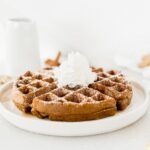 a gingerbread waffle on a white plate topped with powdered sugar and whipped cream.