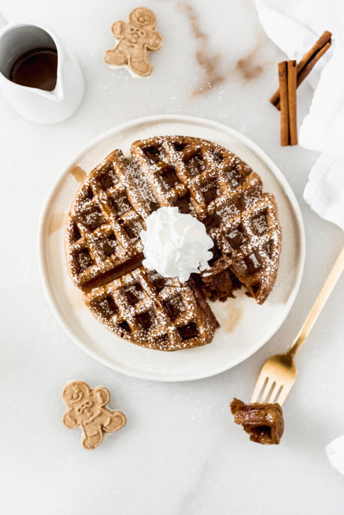 overhead view of a gingerbread waffle on a white plate topped with whipped cream and powdered sugar with a bite taken out surrounded by gingerbread men, cinnamon sticks, and a small container of syrup.
