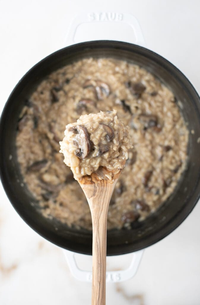 overhead view of brown rice mushroom risotto in a white pot with a wooden spoon lifting some out.