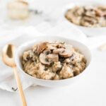 brown rice mushroom risotto in a white bowl with a gold spoon beside it.