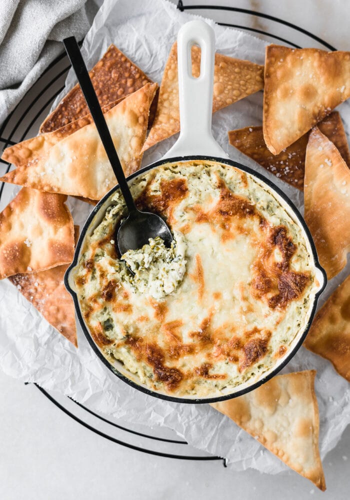 overhead view of healthy kale artichoke dip in a white skillet with a black spoon in it surrounded by baked wonton chips.